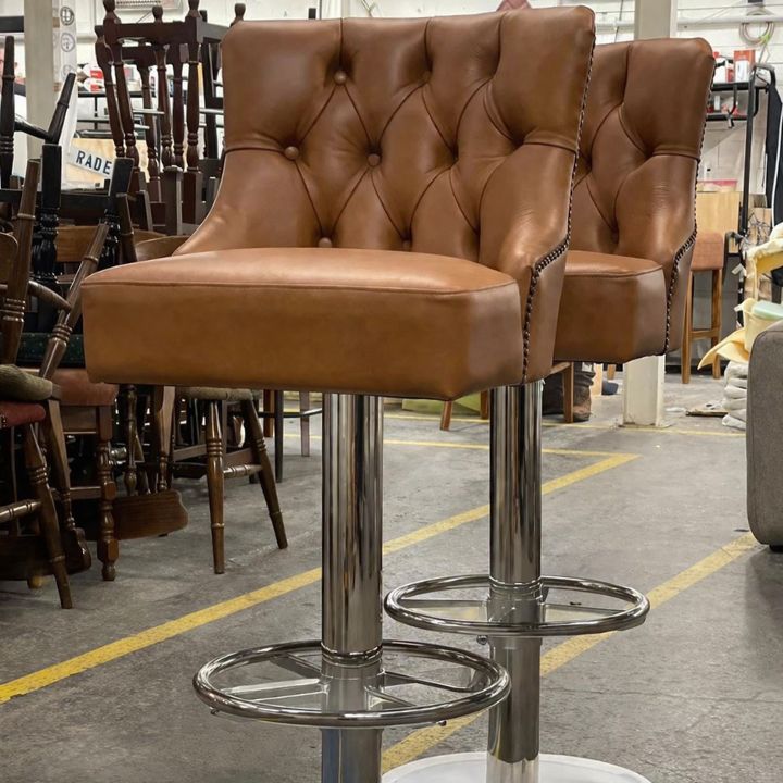 Two brown button-tufted upholstered bar stools with curved backs and chrome pedestal bases, set in a furniture workshop with other chairs and materials in the background.