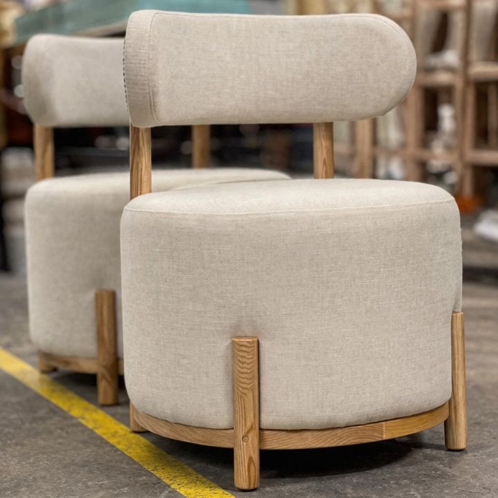 Two beige upholstered barrel chairs with rounded backs, wooden frame accents, and integrated backrests, placed in a furniture workshop with other seating in the background.