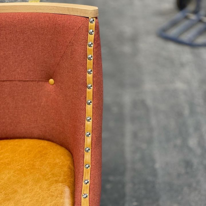 Close-up of an upholstered dining chair with textured fabric back, leather seat, and decorative nailhead trim along a wooden edge.