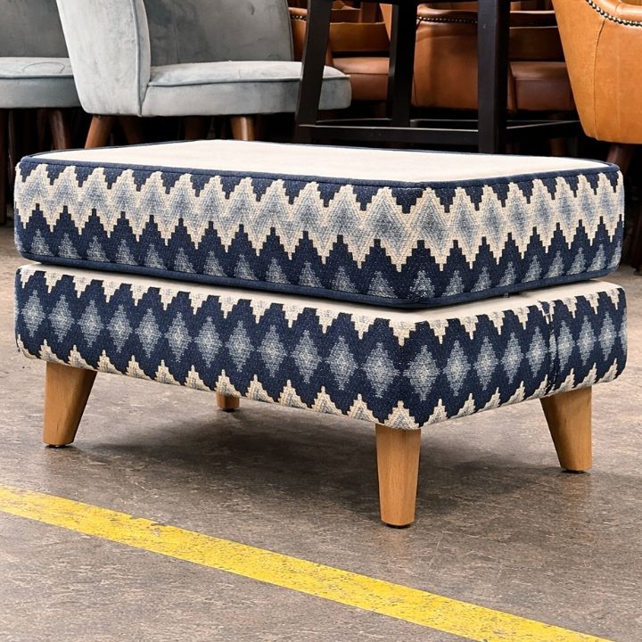 A rectangular upholstered footstool with a blue and white geometric patterned fabric, wooden legs, and a cushioned top, placed in a furniture workshop with chairs in the background.
