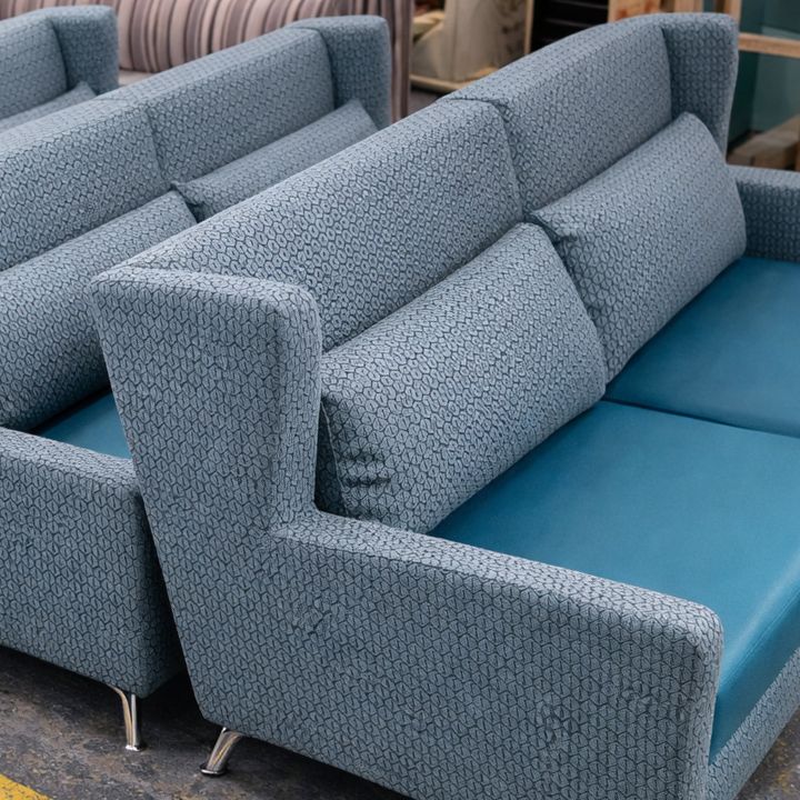 Two blue upholstered lounge sofas with patterned fabric backs, plain seat cushions, and matching scatter cushions, positioned in a furniture workshop with other seating in the background.
