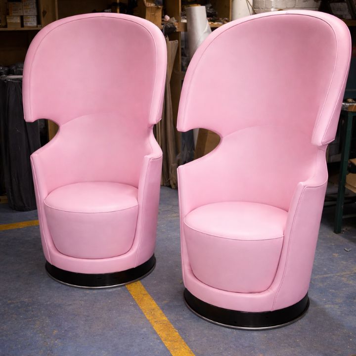 Two pink upholstered pod chairs with high curved backs, enclosed sides, and round black bases, placed in a furniture workshop with materials and tools in the background