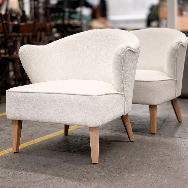 Two cream bouclé upholstered tub chairs with curved backs and light wooden legs, placed in a furniture workshop with other chairs in the background.