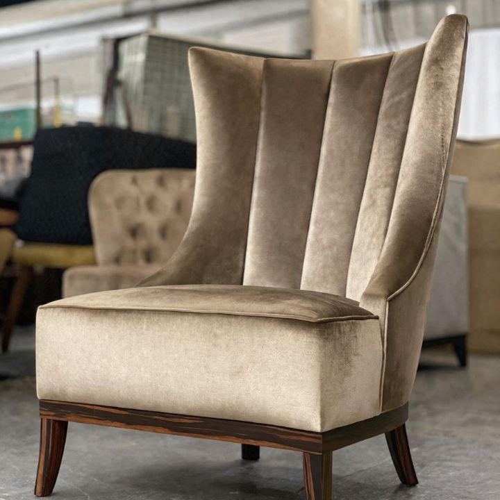 A beige upholstered wingback lounge chair with a high back, vertical channel stitching, and wooden legs, placed in a furniture workshop with other seating in the background.
