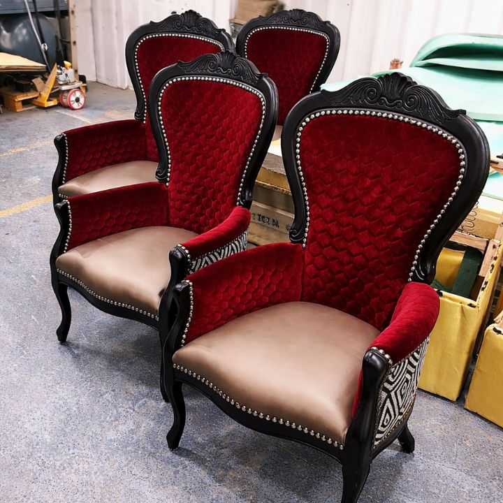 A set of four ornate wingback armchairs with red tufted upholstery, black carved wooden frames, and studded trim, placed in a furniture workshop with materials and equipment in the background.