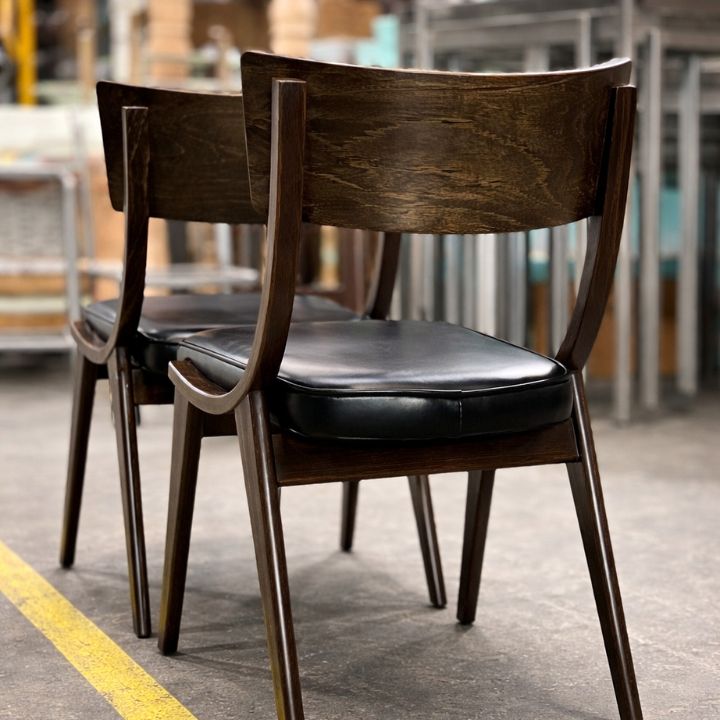 Two dark wooden dining chairs with curved backrests and black upholstered seats, placed in a workshop with furniture and materials in the background.
