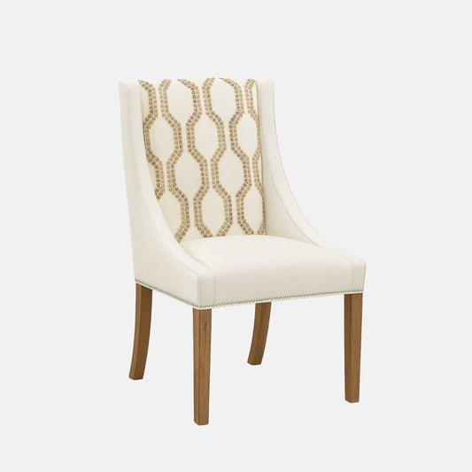 Upholstered dining chair in cream fabric with a gently curved backrest, featuring a geometric patterned fabric panel in beige and gold tones, nailhead trim along the seat base, and natural wooden legs, shown on a white background