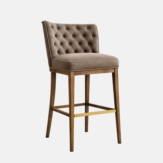 Taupe upholstered bar-height stool with a button-tufted backrest and thick cushioned seat, on a dark wooden four-leg frame with square stretchers and a brass-toned footrest, photographed on a white background.