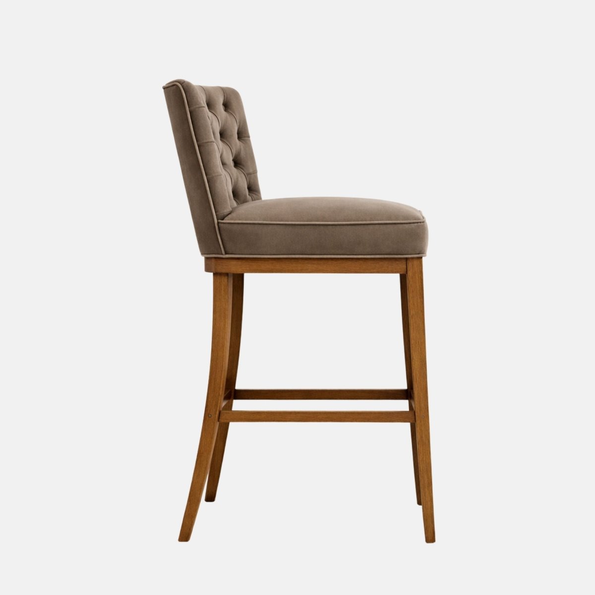 Side view of taupe velvet bar stool with button-tufted back, cushioned seat, and wooden legs with stretcher rails on a white background