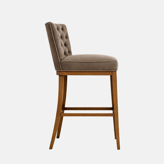 Side view of taupe velvet bar stool with button-tufted back, cushioned seat, and wooden legs with stretcher rails on a white background
