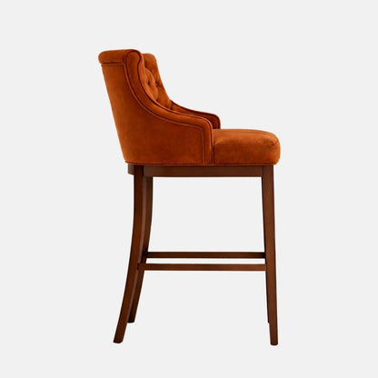 Side profile of a bar-height stool upholstered in warm caramel/cognac velvet or suede-like fabric, featuring a rolled top edge and subtle button-tufting on the inner back, with gently curved arms, a thick cushioned seat, and a dark wood four-leg frame with lower stretchers, photographed on a white background.