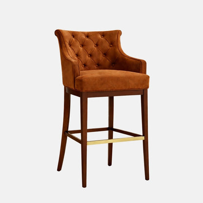 Three-quarter view of a bar-height stool in warm caramel/cognac velvet or suede-like upholstery with a diamond-tufted, buttoned back and gently flared arms, a thick cushioned seat, and a dark wood four-leg frame with square stretchers and a brass/gold foot rail, photographed on a white background.
