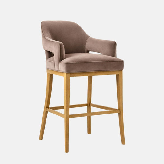 A bar-height upholstered stool in taupe/grey velvet with a wraparound curved back and integrated arms (open cutouts under the arms), a thick cushioned seat, and a light oak wooden frame with four legs and square stretchers, photographed on a white background.
