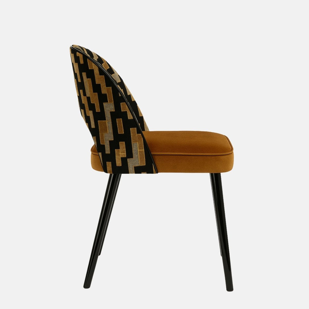Modern upholstered dining chair with a patterned black, gold, and beige fabric backrest featuring a cut-out design, paired with a rust-orange velvet seat and slim black metal legs, set against a light grey background.