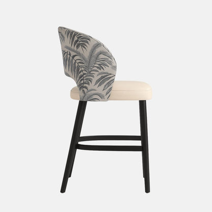 Modern bar stool with curved backrest upholstered in grey fabric with black tropical leaf pattern, cream cushioned seat, and black wooden legs with footrest.