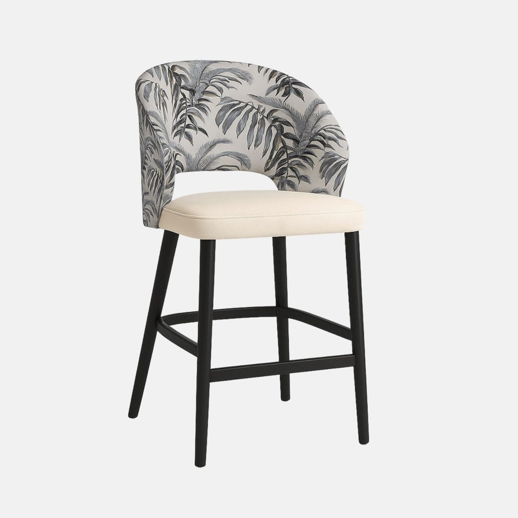 Modern bar stool with curved backrest upholstered in grey fabric with black tropical leaf pattern, cream cushioned seat, and black wooden legs with footrest.