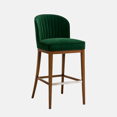 Bar-height stool upholstered in deep green velvet with a curved, vertically ribbed back and thick padded seat, set on a medium-toned wooden frame with four legs, square stretchers, and a metal footrest, photographed on a white background.