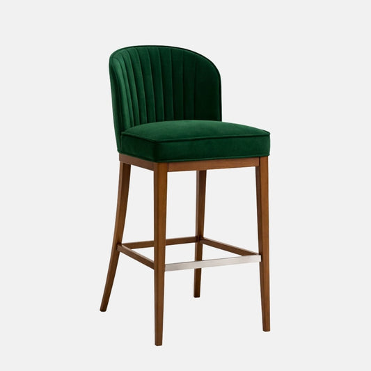 Bar-height stool upholstered in deep green velvet with a curved, vertically ribbed back and thick padded seat, set on a medium-toned wooden frame with four legs, square stretchers, and a metal footrest, photographed on a white background.