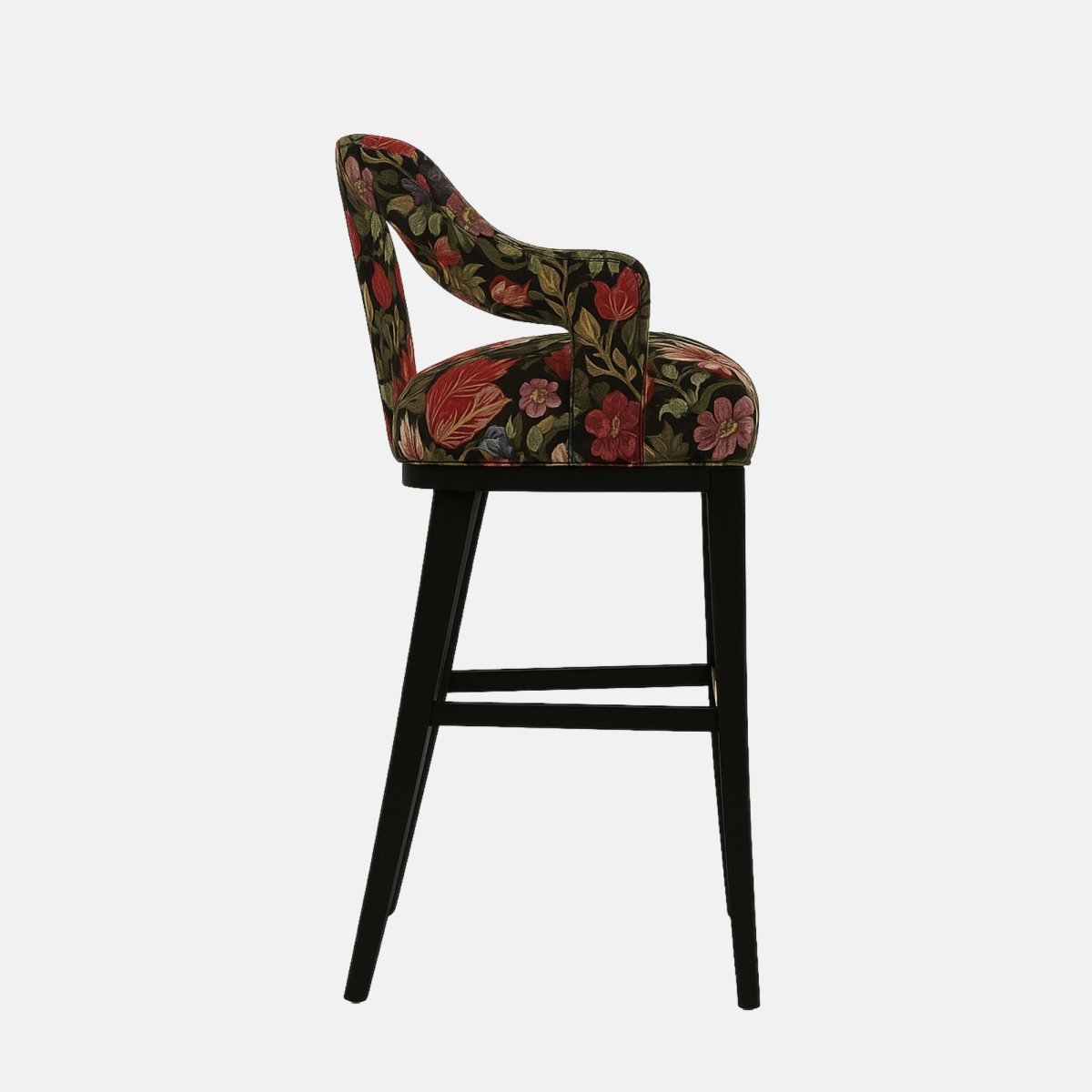Stylish bar stool with a partially open backrest and curved arm supports, upholstered in a bold floral fabric featuring red, green, and gold tones on a black background. It has a black wooden frame with a brass footrest accent, combining vintage and modern aesthetics