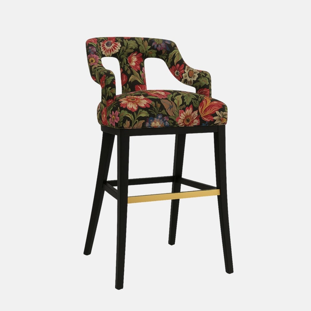 Stylish bar stool with a partially open backrest and curved arm supports, upholstered in a bold floral fabric featuring red, green, and gold tones on a black background. It has a black wooden frame with a brass footrest accent, combining vintage and modern aesthetics