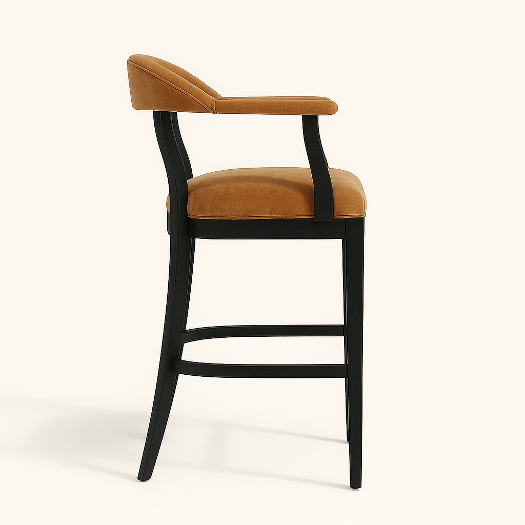 A modern bar stool with a matte black wooden frame and caramel beige velvet upholstery. The chair features curved back legs, a low curved backrest, and padded armrests, all upholstered in matching velvet. The side profile highlights the sleek, ergonomic design and horizontal footrest. The stool is set against a plain off-white background.