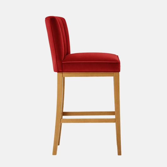 Side view of a red velvet bar stool with a gently rounded backrest, padded seat, and light wood legs with lower stretcher rails.