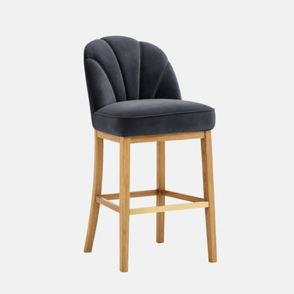 Three-quarter view of a dark charcoal upholstered bar stool with a scalloped, paneled backrest and thick padded seat, on light oak wooden legs with a gold-toned footrest, photographed on a white background.