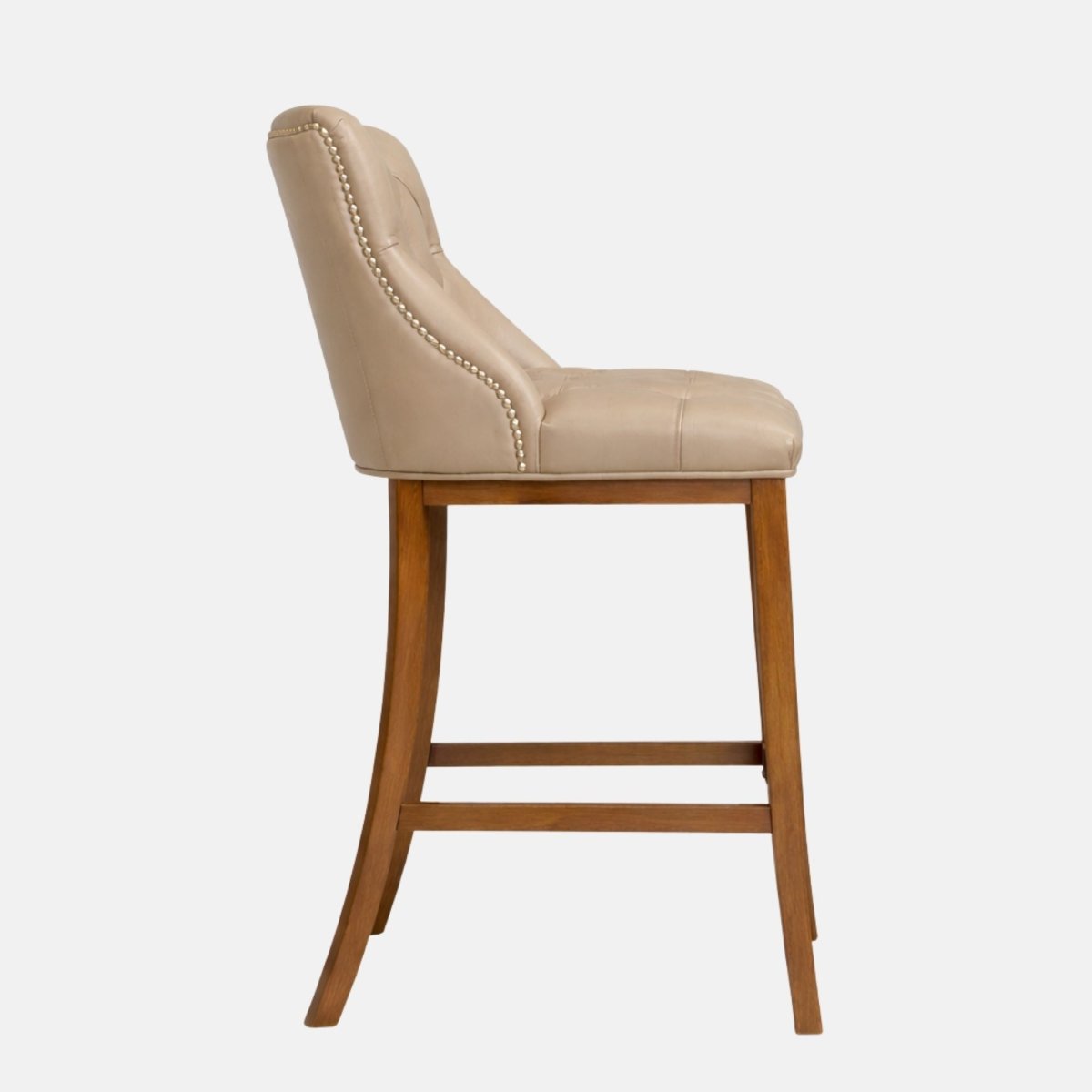 Side profile of a beige upholstered bar stool with a curved, padded back, nailhead trim, and tapered wooden legs with a low stretcher bar.