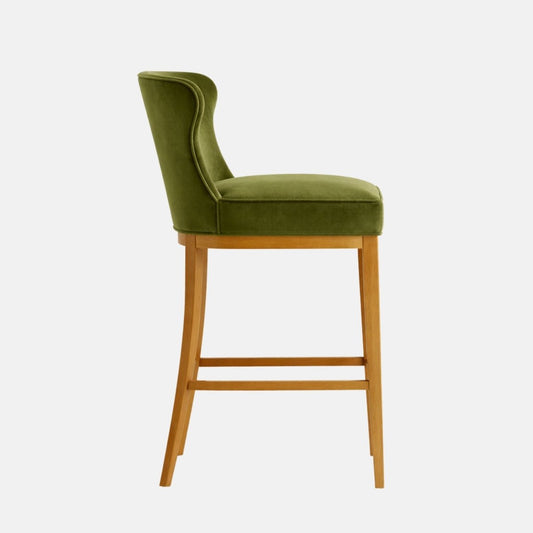 Olive green velvet bar-height stool shown in side profile, with a tall curved back and thick padded seat, on a light oak wooden frame with four tapered legs and two horizontal stretchers, photographed on a white background.