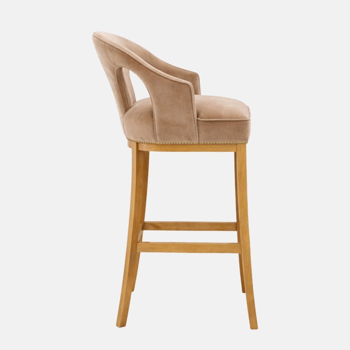 Side view of a taupe upholstered bar stool with curved wraparound back and side cutout, piped seat cushion, nailhead trim, and light wood legs with lower stretcher rails.