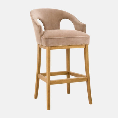 Taupe upholstered bar stool with curved wraparound back and side cutouts, piped seat cushion, nailhead trim, and light wood legs with a square stretcher/footrest.