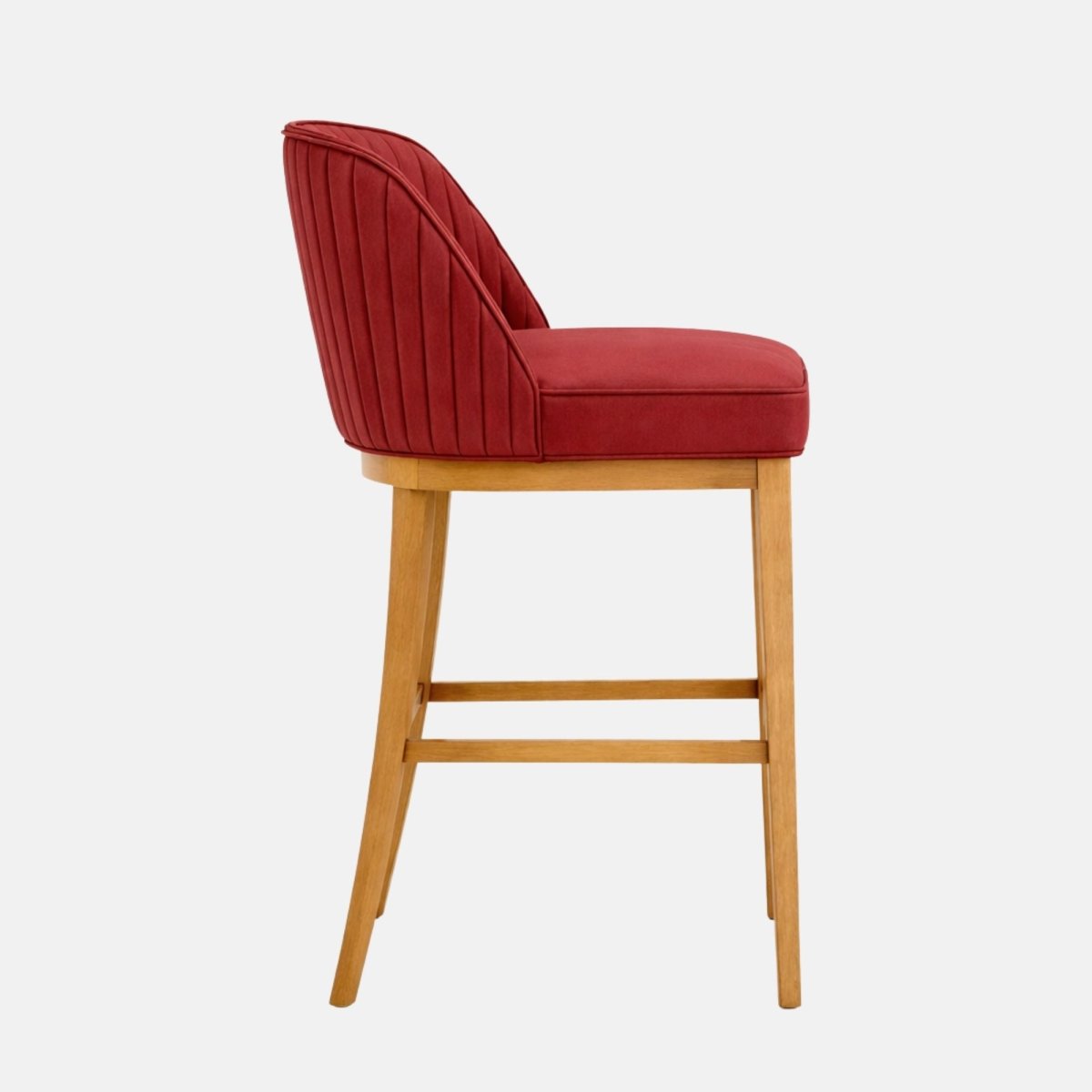 A red upholstered bar stool with a curved, vertically channel-stitched back and cushioned seat, set on a light oak wooden frame with four legs and a square stretcher/footrest, photographed in side view on a white background.