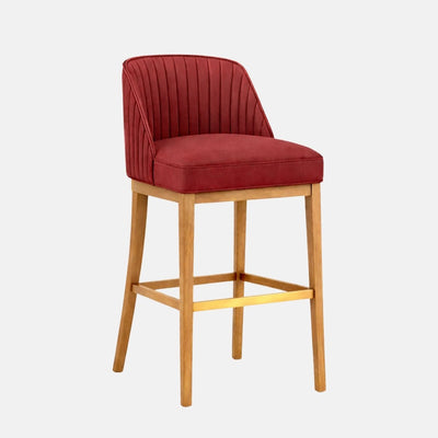 A red upholstered bar stool with a curved, channel-stitched wraparound back and cushioned seat, on a light oak wooden frame with square stretchers and a brass footrest, photographed on a white background.