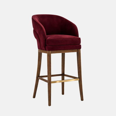 A burgundy velvet bar stool with a curved wraparound tub back and thick cushioned seat, on dark wooden legs with square stretchers and a brass footrest, photographed on a white background.
