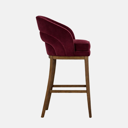 Side view of a burgundy velvet bar stool with a curved wraparound back and thick cushioned seat, on dark wooden legs with square stretchers, photographed on a white background.
