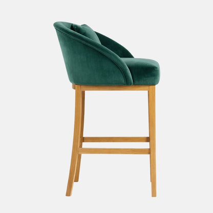 Side view of an emerald green velvet bar stool with a curved wraparound back, padded seat, and light wooden legs with a footrest rail.
