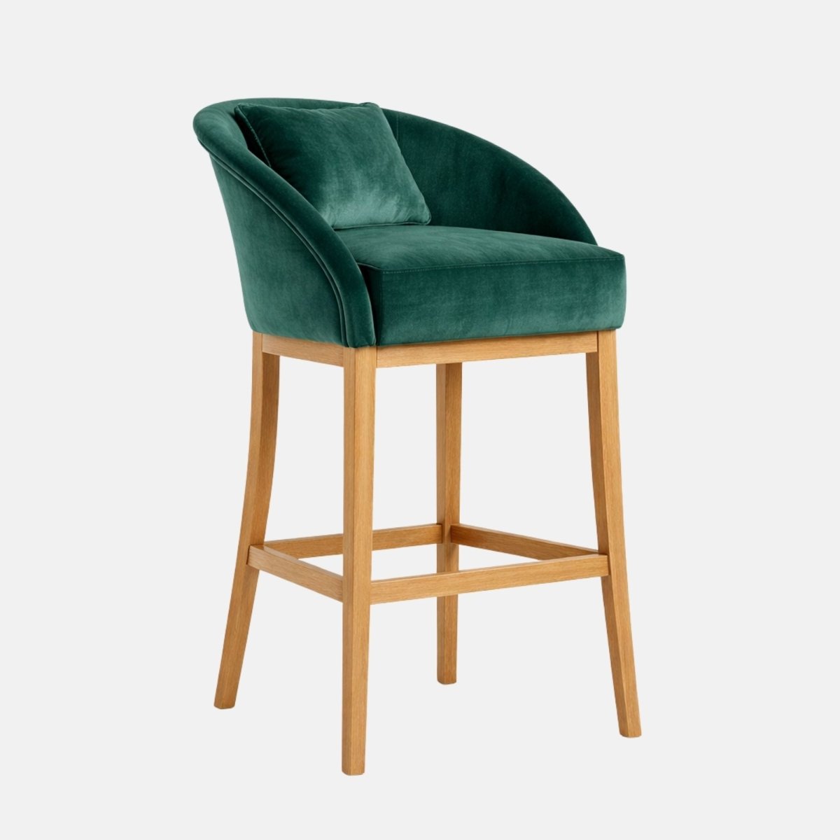 Three-quarter view of an emerald green velvet bar stool with a curved wraparound back, a loose back cushion, a thick padded seat, and light wooden legs with a square footrest frame.