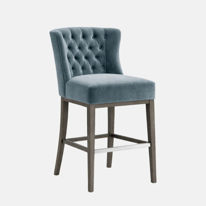 A bar-height wingback stool with a light blue/teal velvet upholstered seat and high tufted backrest, set on a dark grey wooden frame with four legs and a silver metal footrest bar, photographed on a white background.