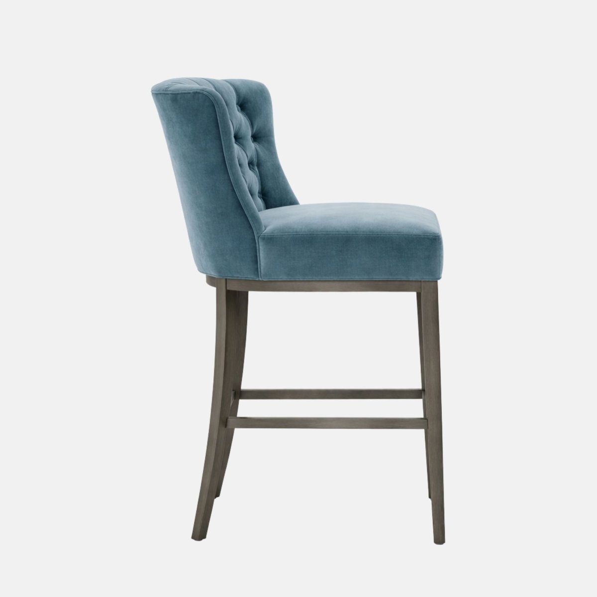 Side view of a bar-height stool with a tall, slightly winged, button-tufted upholstered backrest and a thick padded seat in light blue velvet, set on a dark wood four-leg frame with two horizontal stretcher bars, photographed against a white background.