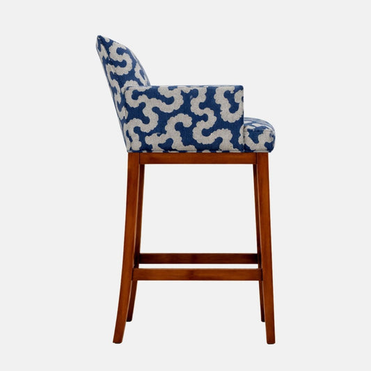 Side view of a blue-and-cream patterned upholstered bar stool with a low wraparound back and arms, thick seat cushion, and dark wooden legs with lower stretcher rails.