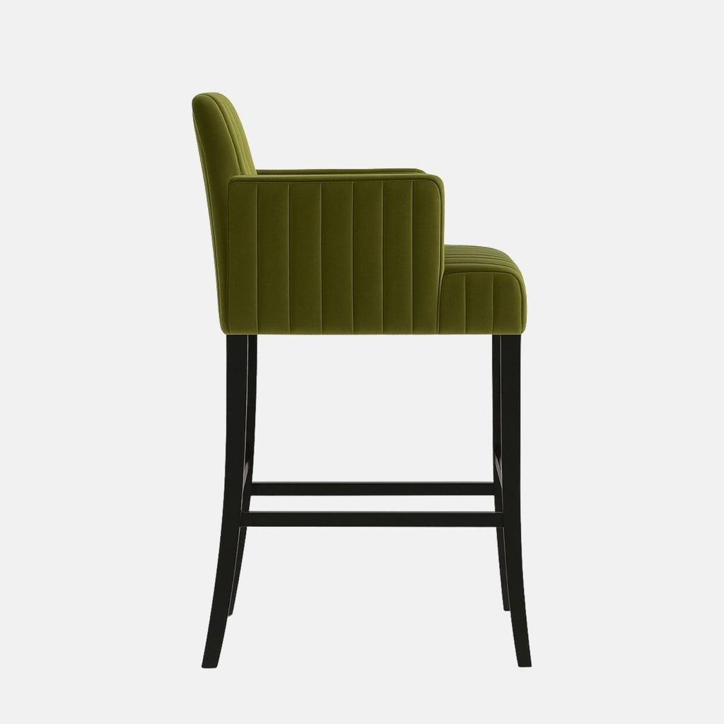 Olive green upholstered bar stool with vertical channel tufting, short armrests, and black wooden legs featuring footrests.