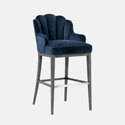 Three-quarter front view of a bar-height stool upholstered in navy blue velvet with a scalloped, channelled wraparound back and integrated arms, a thick cushioned seat, and a grey wooden frame with square stretchers and a silver footrest, photographed on a white background.