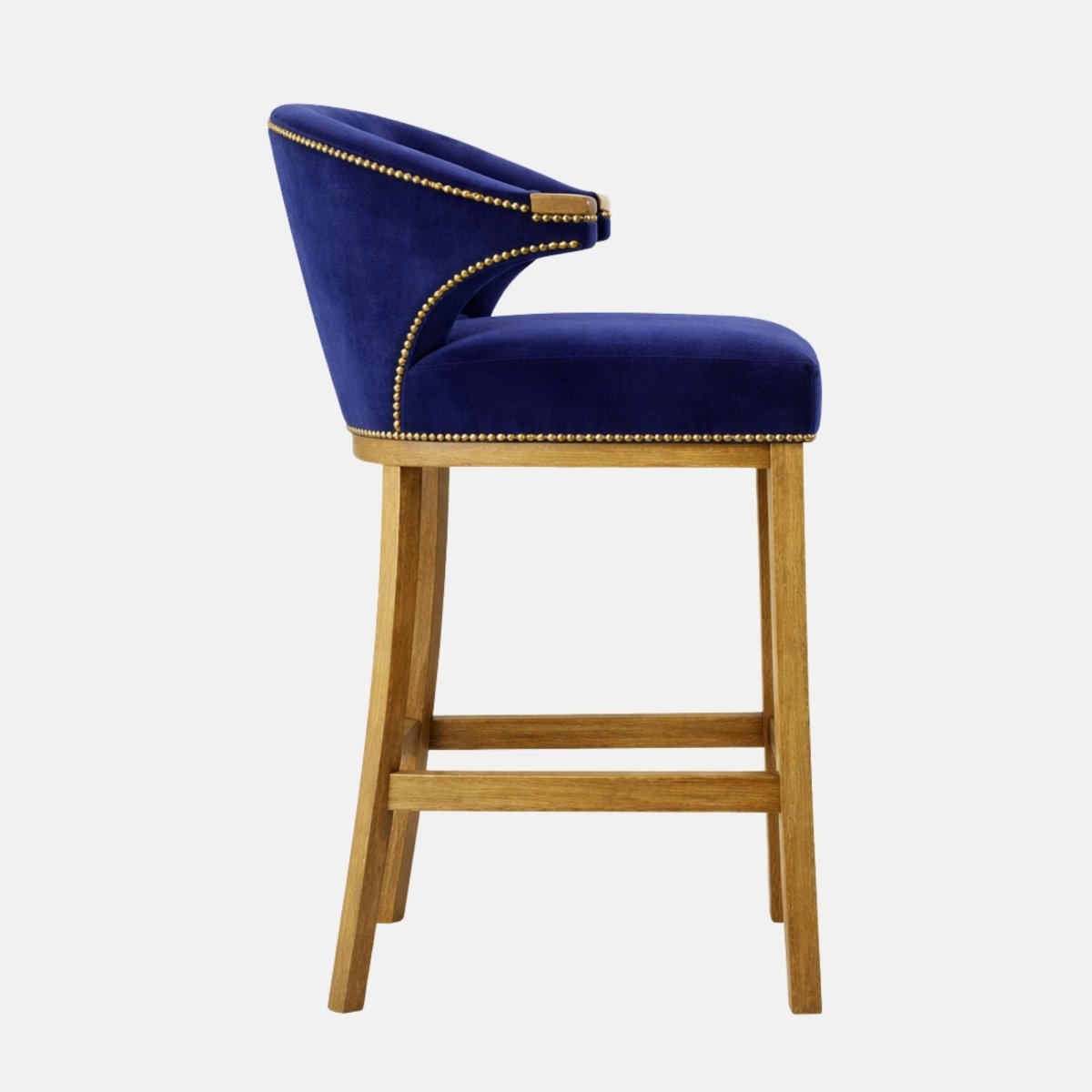Side view of a navy velvet bar stool with a curved wraparound back, nailhead trim, padded seat, and light wood legs with lower stretcher rails.