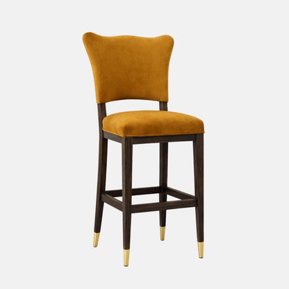Mustard velvet bar stool with a curved backrest, padded seat, dark wooden legs with stretcher rails, and brass leg caps.
