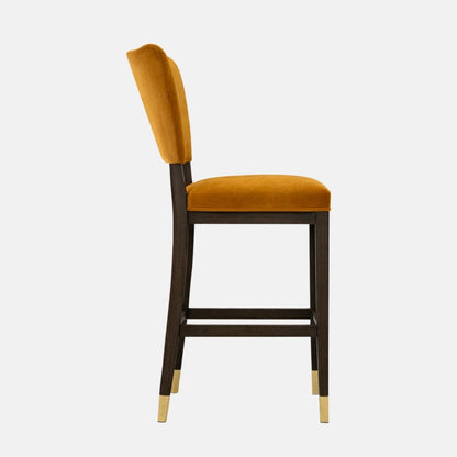 Side view of a mustard velvet bar stool with a curved backrest, padded seat, dark wooden legs with lower stretcher rails, and brass leg caps.