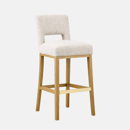 Cream patterned upholstered bar stool with a low backrest and rectangular cutout, padded seat, and light wood legs with a brass footrest bar.
