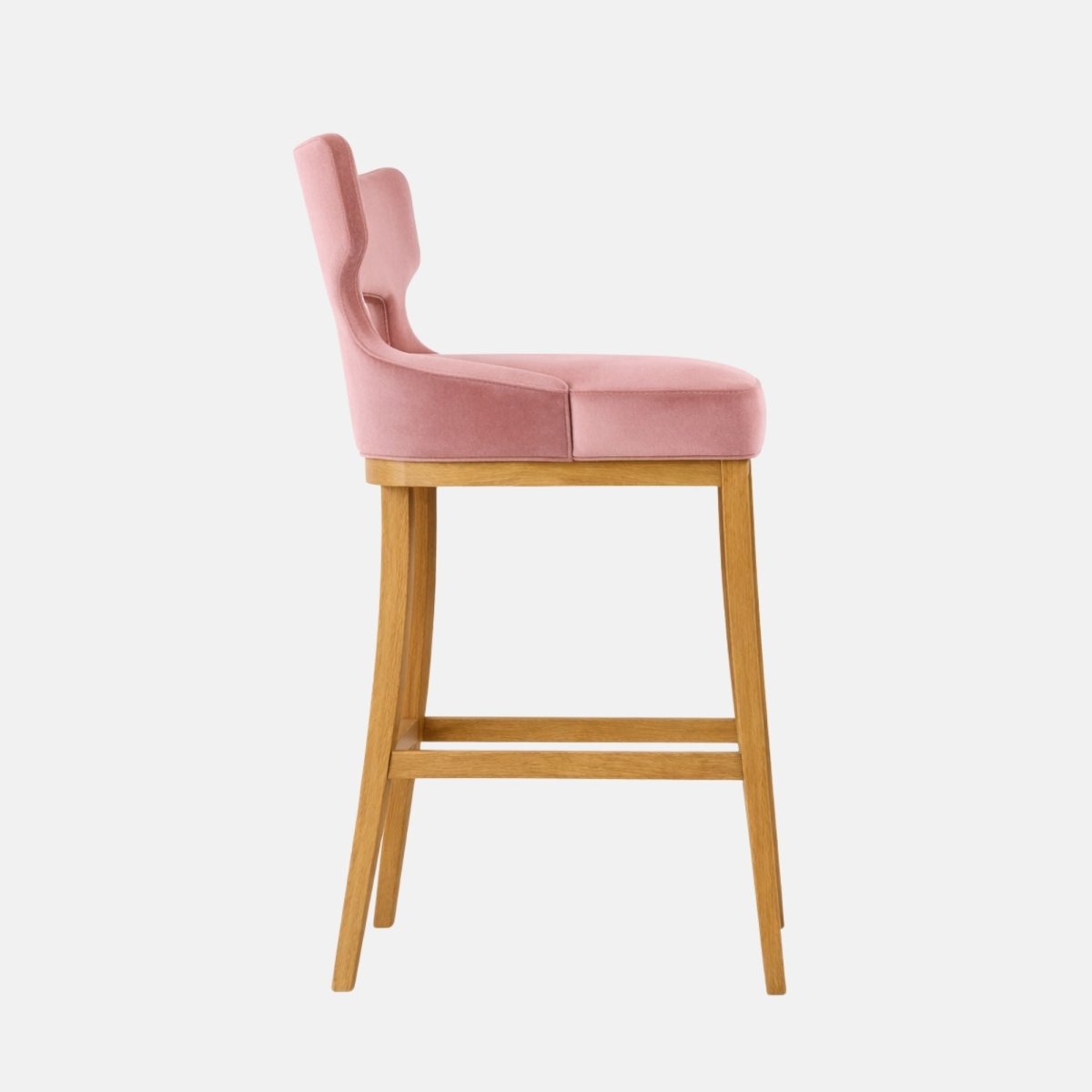 Side view of a bar-height stool with blush pink velvet upholstery and a distinctive open-back/winged backrest silhouette, a thick rectangular padded seat, and a light oak wooden four-leg frame with two horizontal stretchers, photographed against a white background.
