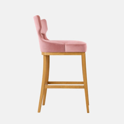Side view of a bar-height stool with blush pink velvet upholstery and a distinctive open-back/winged backrest silhouette, a thick rectangular padded seat, and a light oak wooden four-leg frame with two horizontal stretchers, photographed against a white background.