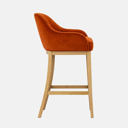 Side view of a burnt orange velvet bar stool with a rounded wraparound back and cushioned seat on light wooden legs with a lower support rail, photographed on a white background.