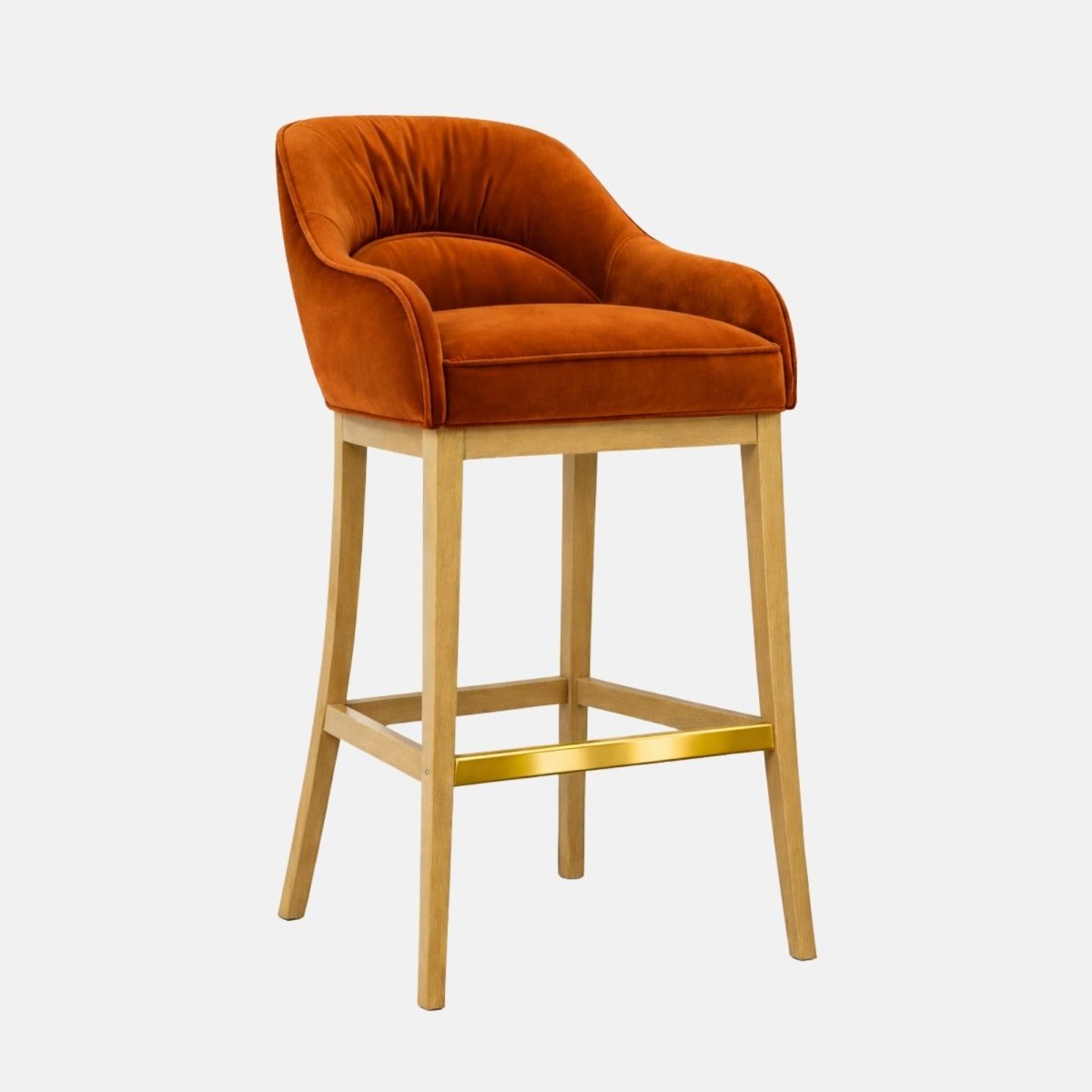 Three-quarter view of a burnt orange velvet bar stool with a curved wraparound back, cushioned seat, light wooden legs, and a gold metal footrest bar.